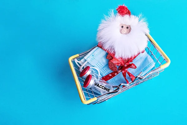Minyatür bir alışveriş sepetinde tıbbi koruyucu maskeler, şenlikli bir kurdele ve mavi arka planda koronavirüs aşısı olan Noel Baba ile bağlanmış. Yeni Yıl ve Noel konsepti.