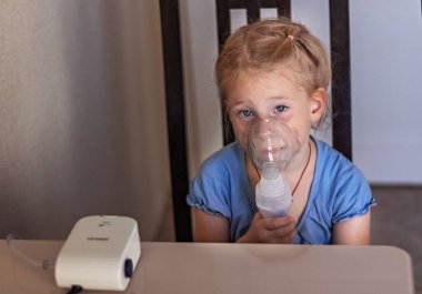 Küçük kız evde bir nebulizörle nefes alıp veriyor, sandalyede oturuyor. Tıkanma ve bronşiyal astım için taşınabilir aygıt..