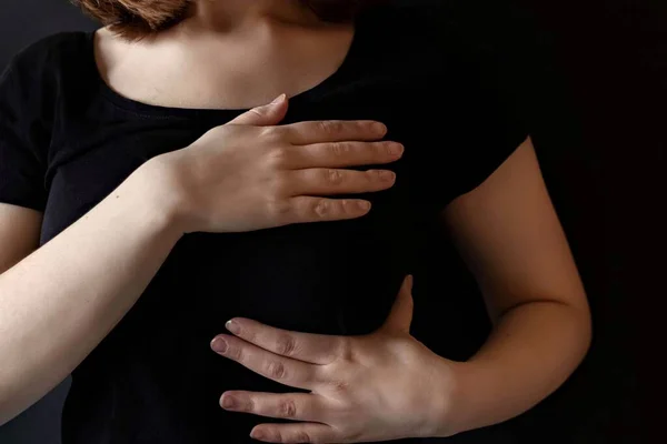 A girl in a black t-shirt clasps her chest with both hands. Women\'s health survey. Breast cancer awareness month. The concept of breast cancer