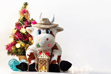 White soft toy bull on a light background with a Christmas tree and garlands, New Year's card. Chinese Year of the Ox, zodiac symbol 2021. clipart