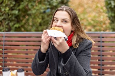 Genç, aç bir kadın hamburger yer ve öğle yemeğini dışarıda dışarıda yer. Fast food. Paket yemek konsepti.