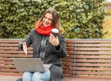 Dizüstü bilgisayarda çalışan genç bir kadın elinde kahveyle parktaki bankta oturuyor. Bilgisayar ve online alışveriş kullanımı. Uzaklık öğrenimi, çevrimiçi eğitim