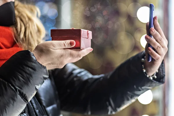Noel Baba şapkalı uzun saçlı bir kadın video iletişimiyle akıllı telefondan iletişim kuruyor. Noel 'de akrabalarını tebrik eder ve bir hediye gösterir. Yeni yıl sokakları. Koronavirüs salgınında sosyal uzaklık.