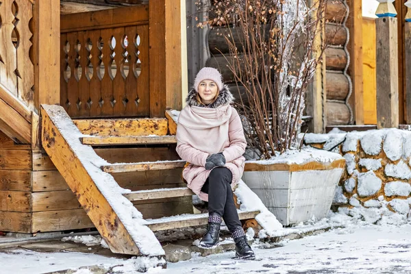Köydeki ahşap bir evin verandasında poz veren sıcacık giyimli genç bir kadın. Kırsalda kış tatili.