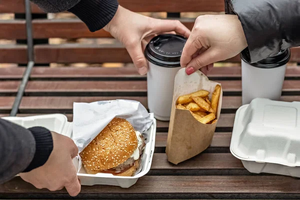 Female hands lay out hamburgers with cups of coffee, fries on a park bench. Fast food. Takeaway food concept.