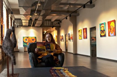 Sanat galerisinin büyük salonunda bir sandalyede oturan genç bir kadın bir kitapçığa bakıyor..