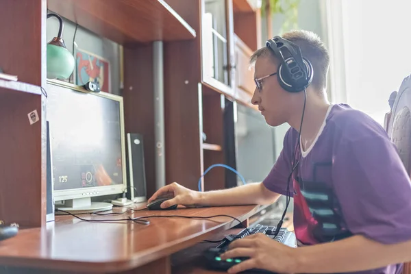 一个少年 一个在计算机上玩电子游戏 使用技术 戴耳机 使用计算机的青年男子 一个使用技术的少年 他玩电子游戏 使用耳机 图库照片
