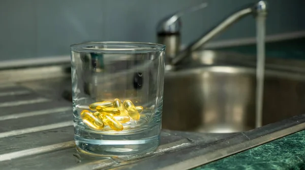 A glass with yellow gold colored capsules  food supplements, vitamins, medicine - placed on a metal sink draining board with running water in the background.