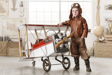 Pilot kıyafetli küçük çocuk odada bir uçakla oynuyor.