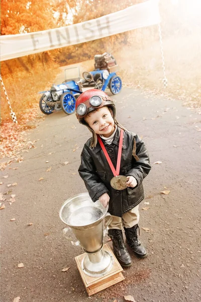 秋の公園で終了時に旧レーサーカーの近くの受賞者の賞を持つレーサーのイメージの小さな男の子 ストック写真