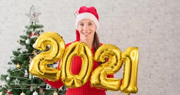 Young woman in Santa Claus hat dancing and having fun holding in hand numbers 2021. Girl in Christmas hat and dancing next to the christmas tree. Concept Christmas celebration girl having fun rejoices — Stock Video