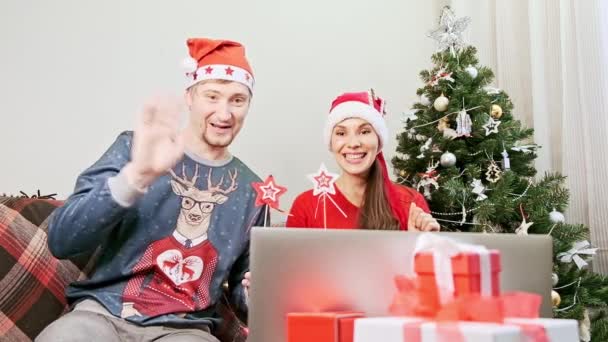 Kvinne med nissehatt og mann kommuniserer med sine kjære på nettet via en bærbar datamaskin. Ung familie gratulerer venner med god jul og godt nytt år under pandemien hjemme foran PC – stockvideo