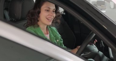 Genç ve güzel bir kız araba galerisinde yeni bir araba seçiyor. Kadın mutlu, gülümsüyor ve yeni bir araba almaktan zevk alıyor. Yeni bir araca saygı duyan bir kız. Alıcı katalogdan bir araba seçer