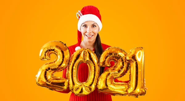 Nouvel An 2021 ballons d'or. Jeune femme en chapeau de Père Noël ayant souriant tenant dans la main 2021. Fille en chapeau rouge et robe rouge se tient sur fond orange vif. Concept Fête de Noël — Photo