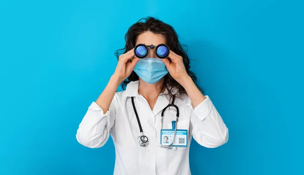 Доктор в маске на ярком цветном фоне смотрит через бинокль. Девушка в концепции защиты covid-19. Баннерное медицинское оборудование для профилактики коронавируса. Женщина в медицинской маске ковид-2019 — стоковое фото