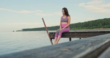 Eşofmanlı genç kız deniz kıyısında iskelede oturur ve bacaklarını sallar. İskelede oturan kadın yaz mevsiminde deniz kenarında dinleniyor.