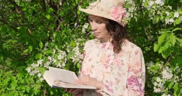 Au ralenti, une fille en chapeau assise dans un parc près d'un arbre à fleurs lit un livre. Gros plan, vue d'une jolie jeune femme souriante lisant un livre et regardant autour dans le parc — Video