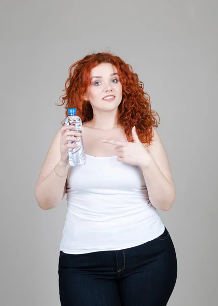 Krásná tlustá dívka s červenými vlasy s lahví minerální vody ruce na šedém pozadí — Stock fotografie