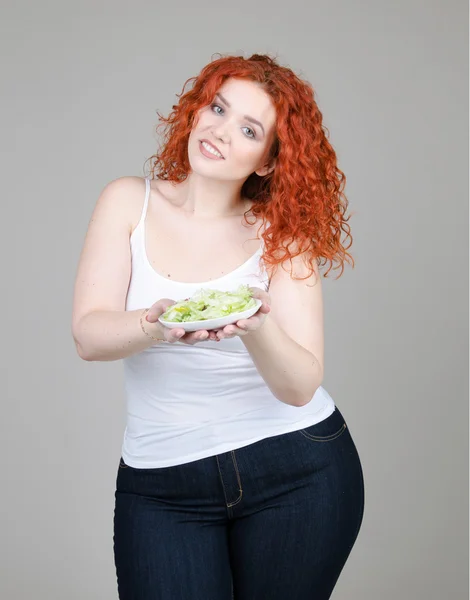 Hermosa chica gorda con el pelo rojo con un plato de ensalada en la mano sobre fondo gris — Foto de Stock