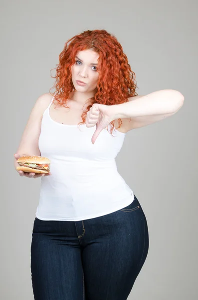 Hermosa chica gorda con el pelo rojo con hamburguesa en la mano sobre fondo gris — Foto de Stock