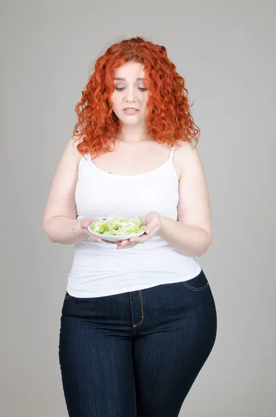 Hermosa chica gorda con el pelo rojo con un plato de ensalada en la mano sobre fondo gris — Foto de Stock