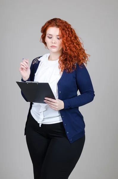 Bella ragazza grassa con i capelli rossi in un abito da lavoro con tablet e penna nelle sue mani su sfondo grigio — Foto Stock