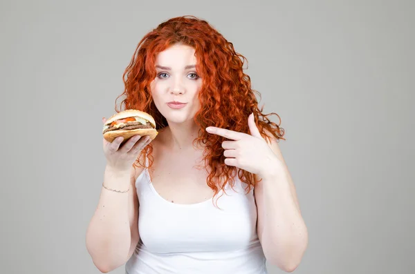 Hermosa chica gorda con el pelo rojo con hamburguesa en la mano sobre fondo gris — Foto de Stock