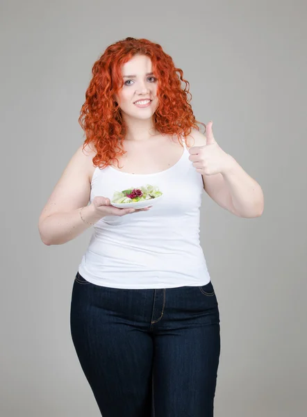 Hermosa chica gorda con el pelo rojo con un plato de ensalada en la mano sobre fondo gris — Foto de Stock