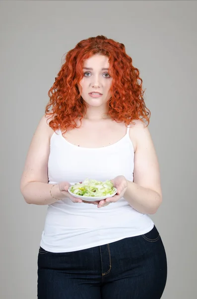 Hermosa chica gorda con el pelo rojo con un plato de ensalada en la mano sobre fondo gris — Foto de Stock