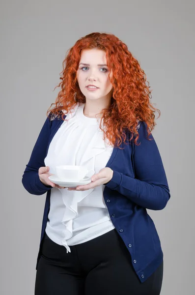 Bela menina gorda com cabelo vermelho em um terno de negócios com uma xícara de chá na mão sobre fundo cinza — Fotografia de Stock