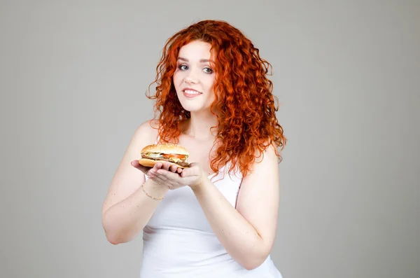 Piękna Gruba dziewczyna z czerwonymi włosami z Burger w ręku na szarym tle — Zdjęcie stockowe
