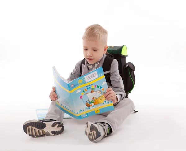 Lindo niño con mochila leyendo un libro en el suelo, aislado sobre fondo blanco — Foto de Stock
