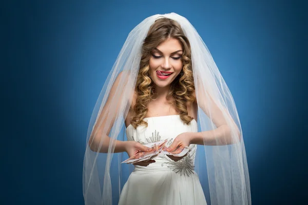 Joven hermosa chica en un vestido de novia con un velo con el dinero en las manos sobre fondo azul —  Fotos de Stock