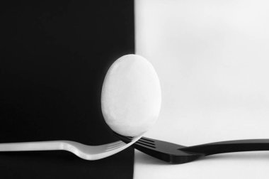 İki birbirine bağlı plastik çatalın üzerinde beyaz bir yumurta duruyor. Arka plan iki parçaya bölünmüş, siyah ve beyaz..