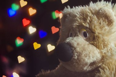Oyuncak ayı, arka planda kalp şeklinde renkli bir bokeh varken kameraya şakacı bir şekilde bakar. Tatil için tebrik kartı..
