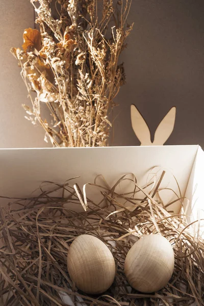 Still life on the theme of Easter. In a box of dried herbs are two eggs behind a box of rabbit ears and a bouquet of dried herbs.