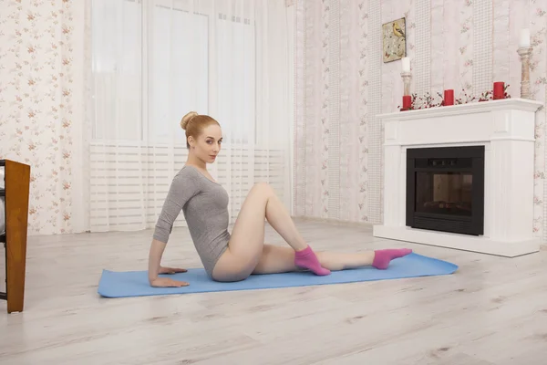 Hermosa mujer rubia practicando yoga estirándose en casa sobre alfombra azul en body gris y calcetines rosados —  Fotos de Stock
