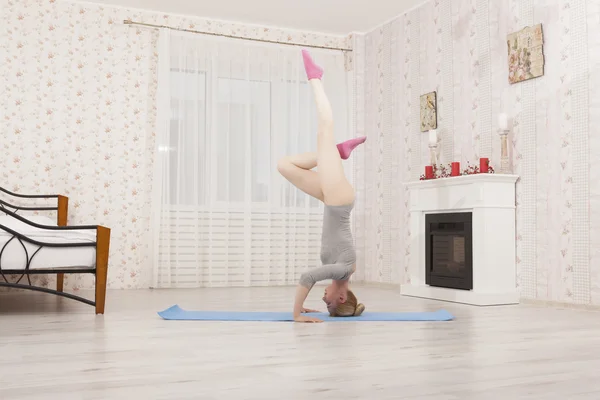 Vacker blond kvinna utövar yoga stretching hemma med blå matta i grå bodysuit, rosa strumpor. Stå på armar och händer — Stockfoto