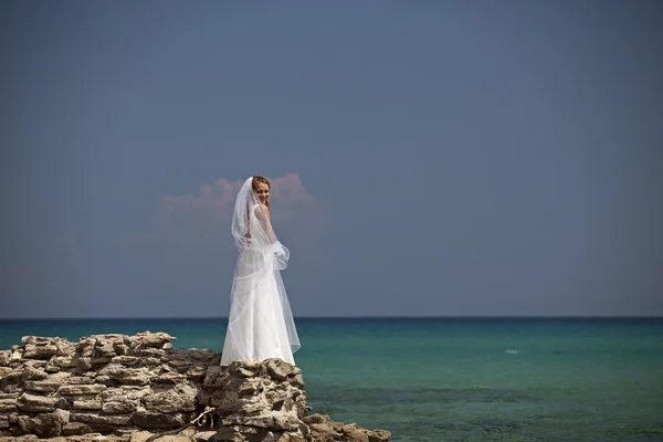 Hermosa novia se encuentra en un acantilado sobre el mar en glamoroso vestido de novia blanco — Foto de Stock