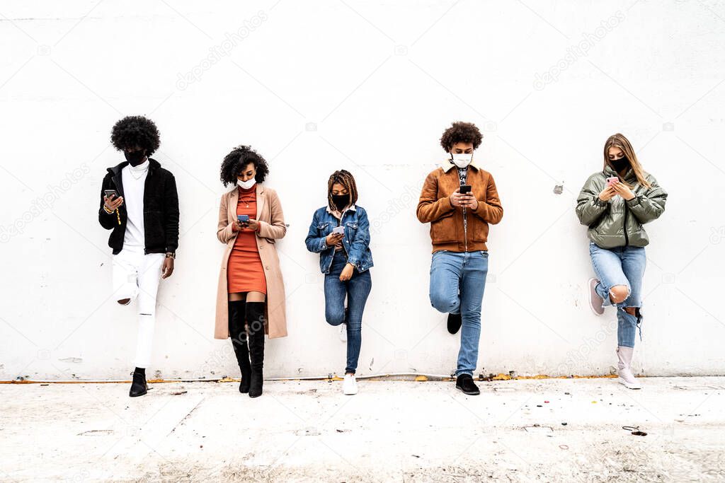 Multiracial group of friends keeping distance outdoor, standing over the wall with face mask covered. Each person is looking at mobile phone and watching new content on social media. 