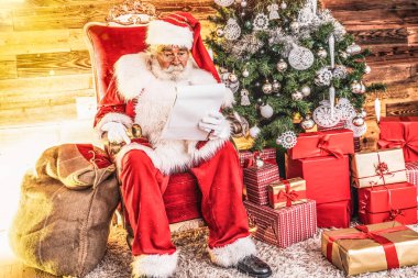 Noel Baba çocukların mektuplarını okuyor, Noel ağacı ve Noel hediyelerinin yanında oturuyor..