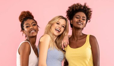 Çok ırklı üç güzel kadının, gülümseyen ve kameraya bakıp pembe pastel arka planda poz verdiği bir moda fotoğrafı. Güzellik yaşam tarzı ve çok ırklı arkadaşlık kavramı. 