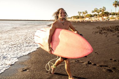 Mutlu yakışıklı adam sahilde sörf tahtasıyla yürüyor. Gülümseyen sörfçü. Hobi olarak. Yaz zamanı. Ada havası. Gerçek insan yaşam tarzı ve ekstrem spor konsepti