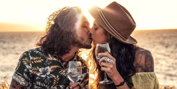 Fashionable boho couple with wine having romantic date on sunset beach, kissing, spending time together. Real people emotions and love concept.