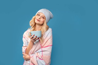 Kışın şık şapkalı ve eşarplı güzel bir sarışın kadın kameraya gülümsüyor, sıcak çay içiyor, mavi stüdyo arka planında izole edilmiş. Gerçek insanların duyguları. Bir sürü kopyalama alanı.