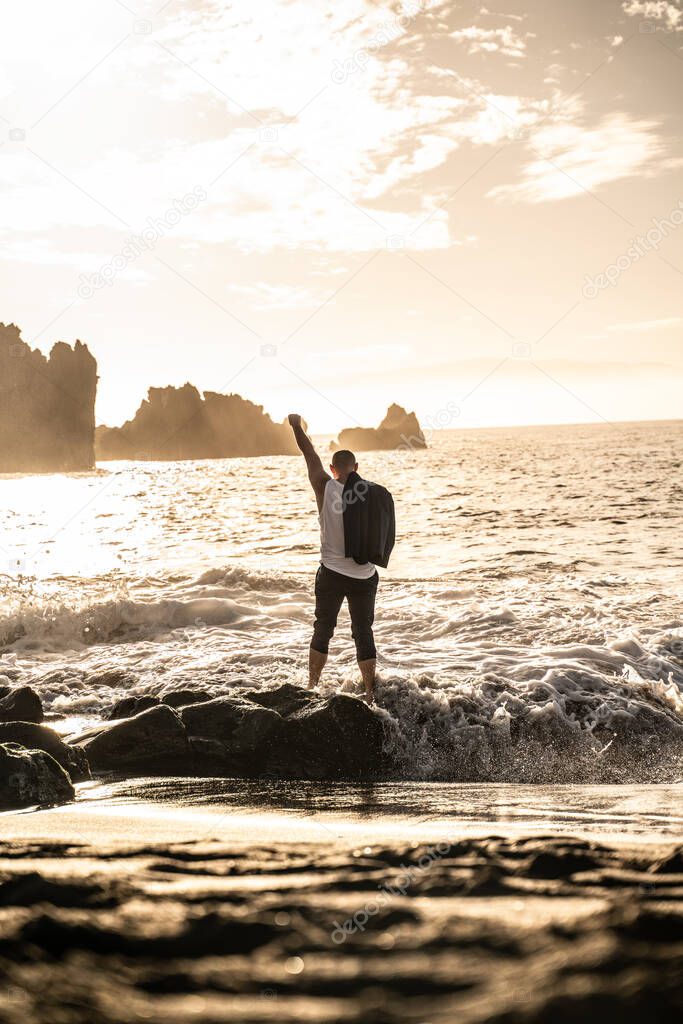 Businessman standing on the rocky coast of the ocean during beautiful sunset with hands up, celebrating success.