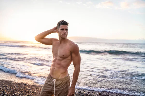 Handsome young muscular italian man posing shirtless on the beach, sunset summer time. Ideal fit body.