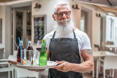 Kahvecinin ya da restoranın önünde gülümseyen bir garsonun gülümseyerek müşterilere içki servisi yaptığı fotoğraf. Beyaz sakallı ve bıyıklı yakışıklı bir son sınıf öğrencisi. İşte mutlu bir işçi..