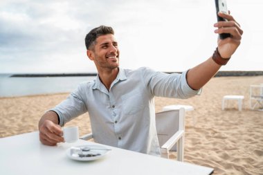 Yakışıklı gülümseyen adam, plaj barında kahve içerek selfie çekiyor. Rahatlıyor, gülümsüyor. Kameralı cep telefonuyla arkadaşlarıyla konuşan bir adam. Sosyal medya saçmalıkları. Etkileyici.