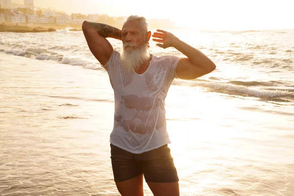 Age is just a number. In a healthy body, healthy mind. Senior man with white stylish beard and muscular fit body with tattoos on the beach.  Sunset light. Elderly people healthy lifestyle on a retirement concept